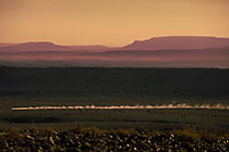 car driving fast w dust on track through rocky desert + mts in haze at sunrise